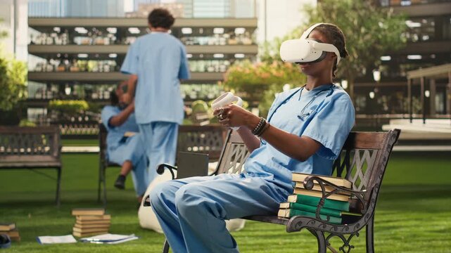 The medical student uses VR controllers and glasses in blue scrubs on a bench, using virtual reality technology for medical training on campus, during an immersive learning study session.