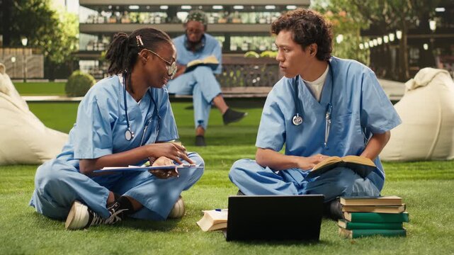 Med school classmates share a fist bump beside a laptop and books on a campus lawn, with friends celebrating a successful group study session.