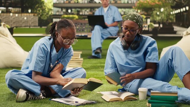 Women medical students in blue scrubs compare open textbooks, discussing coursework on a modern campus lawn, group study notes and academic materials nearby.