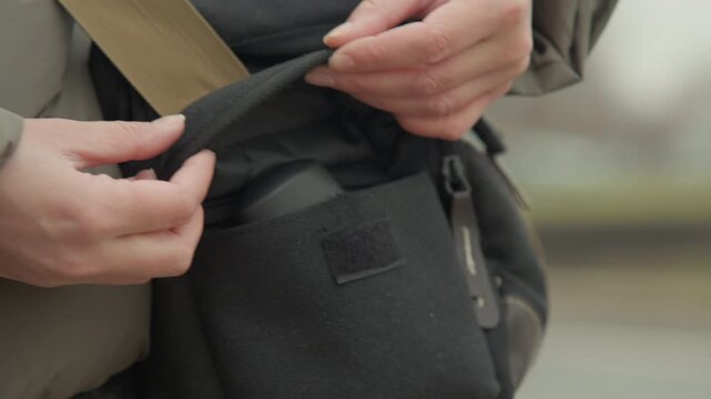Hands opening black canvas pouch outdoors, closeup showing gentle finger movement on flap, khaki strap, textured fabric, muted city background and overcast light, practical retrieval of small camera