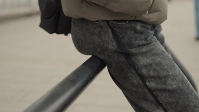 Closeup legs winter pants leaning railing, commuter resting on waterfront boardwalk grey puffer coat, textured knit trousers, subtle frost pattern on fabric, cold overcast light, shallow depth