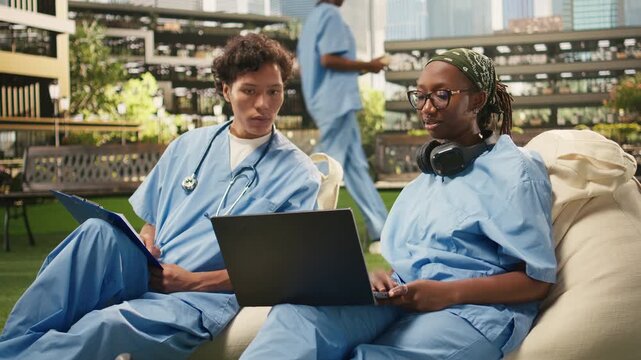 Medical students in blue scrubs compare notes during a relaxed outdoor group study, sitting on beanbags and discussing healthcare, with a laptop and open book serving as study materials.