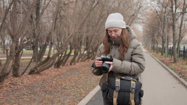 Young woman photographer prepares camera bag on winter park path, pulling mirrorless camera from padded case, adjusting lens and strap, checking settings on screen, wearing beanie and puffer coat,