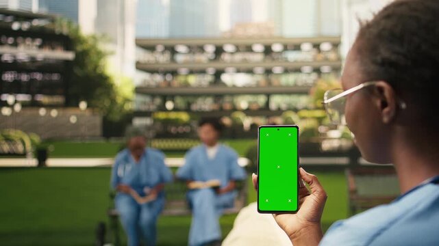 The student's hand holds a smartphone with a vertical green screen mockup in the lower frame, leaving generous copy space over softly blurred med school campus background, during outdoor learning.
