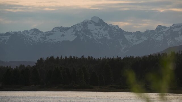 Scenic landscape view of snow-capped mountain peaks rising above a dense pine forest, reflected in the calm waters of Laguna La Zeta in Esquel, Chubut, Argentina.