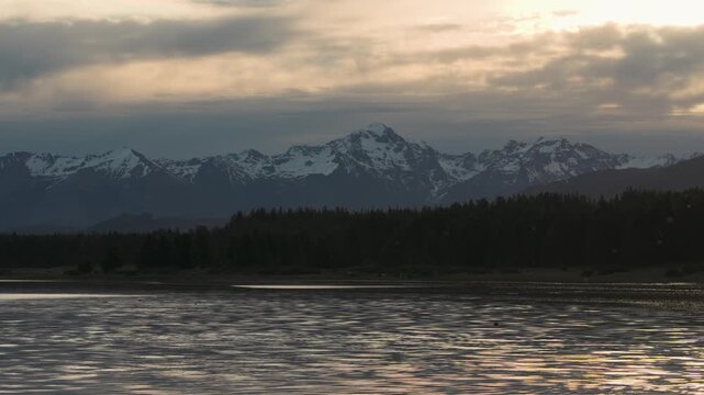 Panoramic view of a rugged mountain range with snow-covered peaks overlooking a calm lake at dusk.