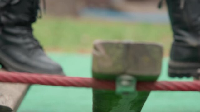 Closeup plank and red rope on low bridge, rugged boot making measured contact with wood, visible bolt and rope tension, textured grain and green safety mat, careful pace and focused inspection
