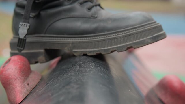 Boots measured steps along textured pipe, precision walker moving slowly over worn tube with red anchors, intimate closeup on sole and knob, quiet reflective mood emphasizing grip, patience