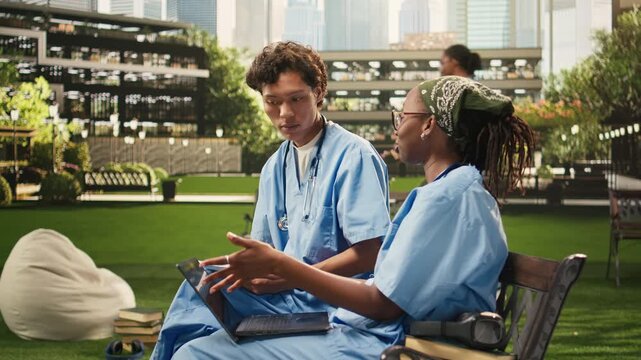 Diverse students in blue scrubs compare notes in an open textbook, discussing coursework during a peer tutoring session on campus bench, with academic materials supporting group study.