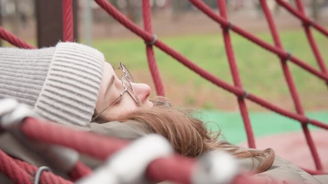 Woman lying on red rope net with knit beanie and glasses, closeup portrait showing soft contemplative gaze and textured lattice framing face autumn park backdrop, muted tones and cozy jacket convey
