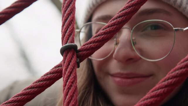 Closeup white girl kissing through rope net, winter beanie and glasses, soft smile and subtle pout, student poet pressing lips toward camera between red ropes, candid cozy portrait in overcast park