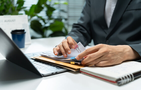 Businessman organizes clipped documents beside a laptop and notebook at an office desk, representing paperwork management, document review, administration, and organized corporate workflow.