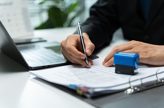 Businessman completes a printed form with a pen beside a laptop and office stamp, representing documentation, application processing, record keeping, approval, and administrative work.