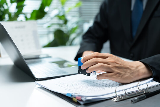 Businessman applies an office stamp to paperwork beside a laptop, representing document approval, verification, authorization, compliance, certification, and administrative processing.