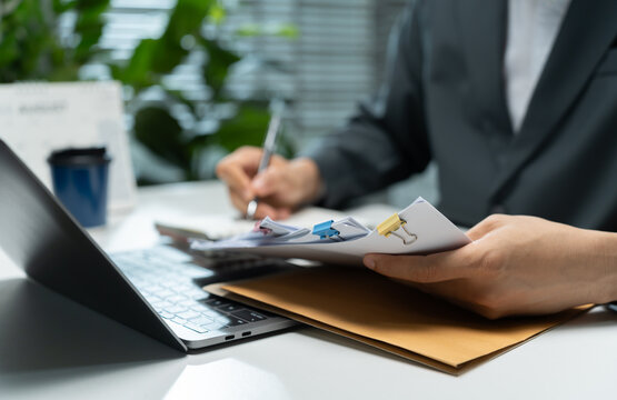Office professional reviews a stack of clipped paperwork beside a laptop while taking notes, illustrating document processing, record checking, administrative tasks, and business office workflow.