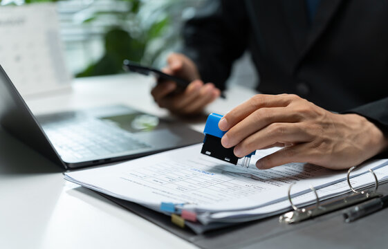Businessman stamps a printed document at an office desk beside a laptop, symbolizing approval, verification, authorization, paperwork processing, compliance, and official administration.