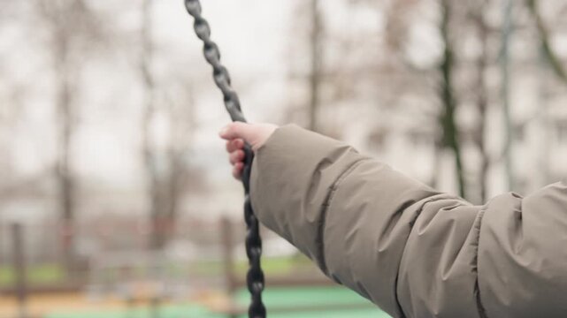 Woman hand gripping metal chain closeup, puffer sleeve and knitted cuff, focused hold and firm tension, textured links and blurred park background, cold season detail shot of playground equipment
