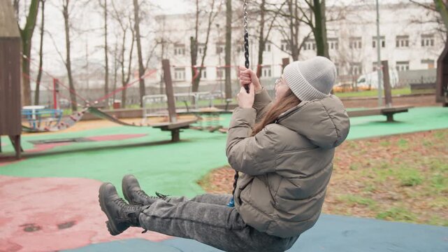 Woman on zipline swing in park wearing puffy jacket and knit beanie, laughing while holding phone, wooden play structure and bare trees under overcast sky, dynamic motion and carefree expression