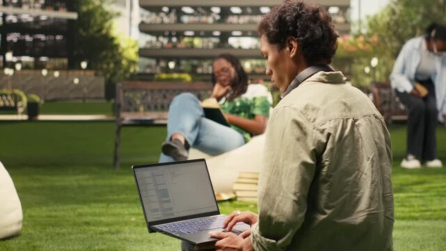 The international student uses a laptop with coding content on a campus bench while classmates study in the background for outdoor learning, for coding and automation study in an academic environment.