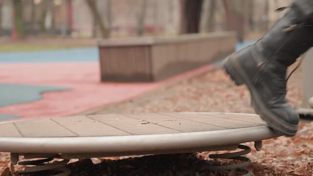 Boots stepping onto spring platform, autumn leaves crunch underboard as lone person tests balance on circular spring pad at neighborhood playground, soft overcast light and muted color palette,
