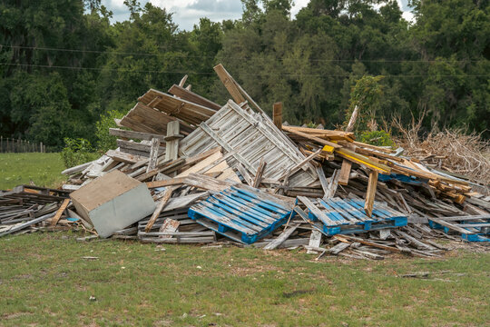 Large pile of discarded wooden shipping pallets and broken wood boards lying on the green field grass near green forest trees outdoors.