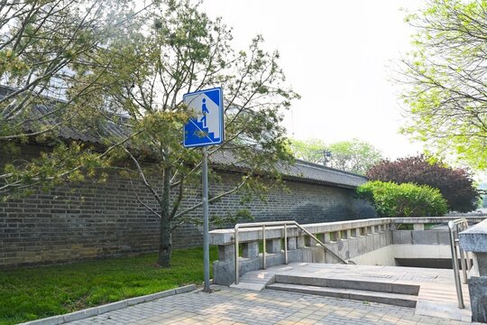 Pedestrian underpass entrance with blue road sign near historical grey brick wall and green trees in street of Beijing China