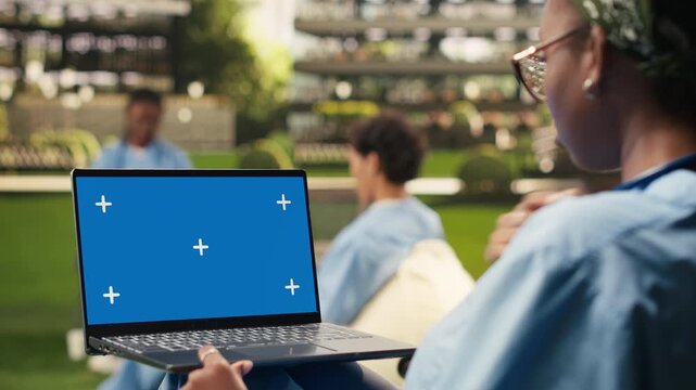 The student holds a laptop with a blue screen mockup and uses the touchpad with one hand, showing an isolated chroma key display ready for replacement in a medical school courtyard.