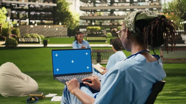The seated medical student holds a laptop with a blue screen mockup on campus, providing an editable chroma key display for computer screen replacement in education content for medical school use.