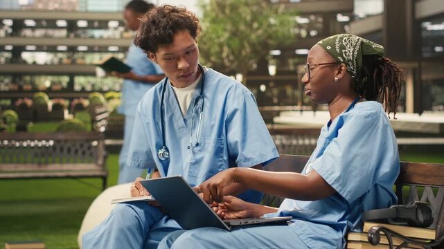 Medical students discuss coursework with a laptop and study materials, sitting on a bench near stacked books on a modern campus, with classmates in a group study session, during peer tutoring.