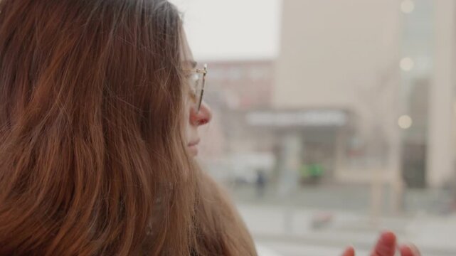 Lady in winter observing snowfall. Female with glasses observing winter snowfall. Brown haired woman wearing glasses stands on city street observing gently falling snow in winter conditions
