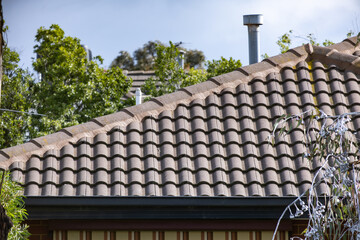 Obraz premium Terracotta tiled roofline of a suburban home with a metal flue vent, guttering, and mature trees in the background. Roof of a residential house in Melbourne, Australia. Concept of roofing, maintenance