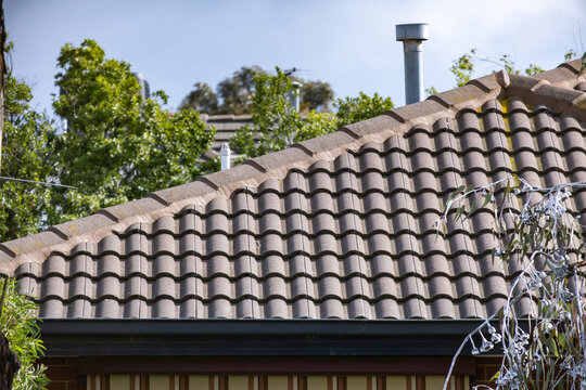 Terracotta tiled roofline of a suburban home with a metal flue vent, guttering, and mature trees in the background. Roof of a residential house in Melbourne, Australia. Concept of roofing, maintenance