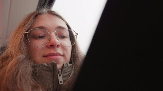 Smiling white woman on tram browsing phone and scrolling messages, wearing glasses and cozy jacket lowangle candid portrait with warm morning light, freelance creator energy and casual commuter