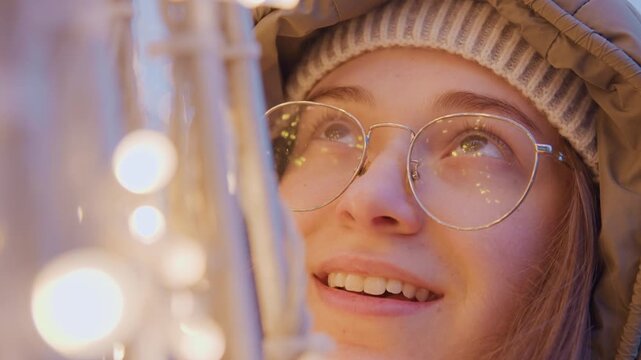 Smiling young woman under festive lights with warm glow, cozy beanie and hood, reflective glasses catching twinkle, gentle gaze, optimistic mood, intimate portrait vibe, street backdrop