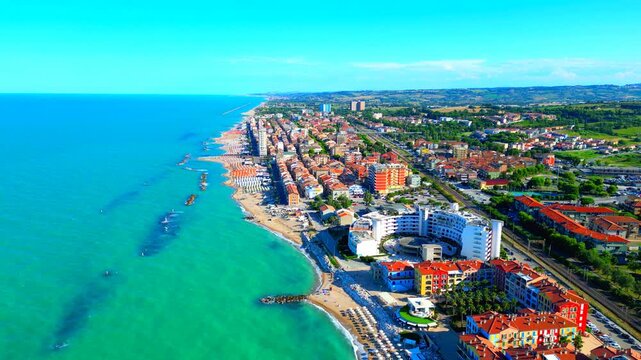 Fantastic aerial pullback truck left pan left shot from Porto Recanati, the Adriatic Sea's emerald waters splashing white foam by the sandy beach, complete with parasols, apartment complexes, houses