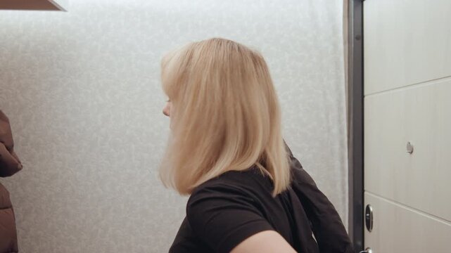 White woman in doorway putting on coat, side profile movement, blonde bob tucking hair, sliding into padded jacket, brief glance downward, everyday departure routine in compact apartment entry, candid