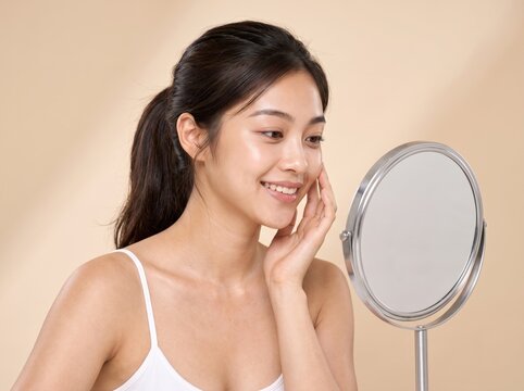 Smiling Asian woman looking in vanity mirror on beige background