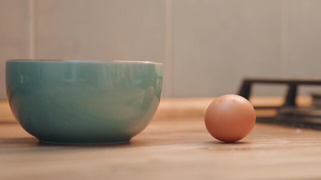 Hand placing egg into ceramic bowl, warm wooden counter, gas stove backdrop, gentle motion of hand setting brown egg beside bowl, closeup domestic scene, breakfast preparation, tactile texture
