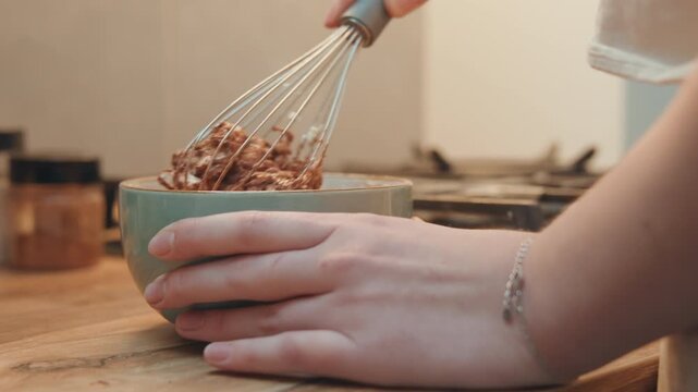 Closeup hands whisking chocolate batter in ceramic bowl on wooden counter, spice jars blurred, soft morning light, home baker mixing slowly with tactile motion and warm atmosphere
