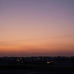 Fototapeta premium city skyline silhouette at dusk seen from a rooftop, large empty sky with orange and purple gradient, a few distant buildings with warm glowing lights
