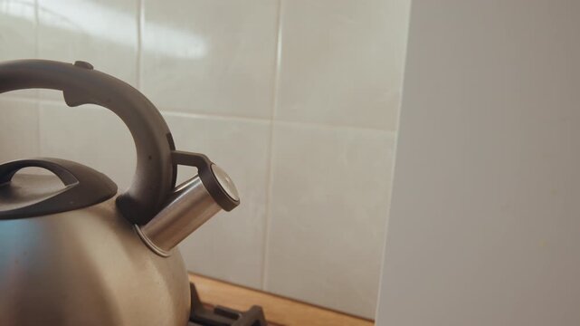 Metal kettle on gas stove, quiet kitchen morning. Stainless kettle sits on lit burner next to tiled backsplash, subtle steam forming, warm ambient light, domestic cooking scene, breakfast routine,