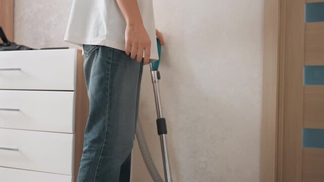 Person vacuuming home hallway floor, canister vacuum blue body, moving along baseboard and dresser, casual jeans white shirt, natural light, quiet morning cleaning routine, focused household chore