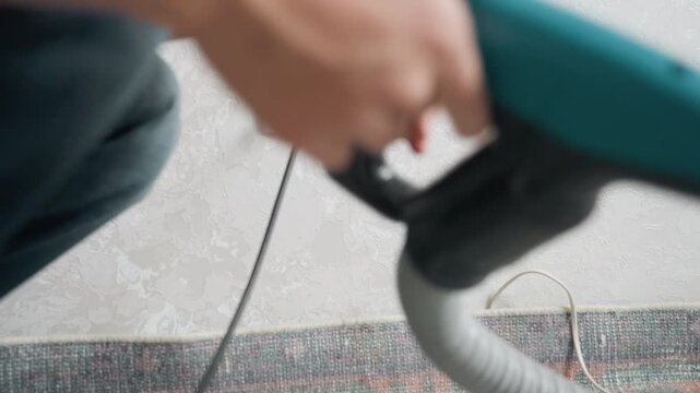 Asian man plugging vacuum into outlet before home cleaning, crouched on carpet with hand gripping plug and hose, careful connection and inspection of cord and socket, closeup on switch and handle,