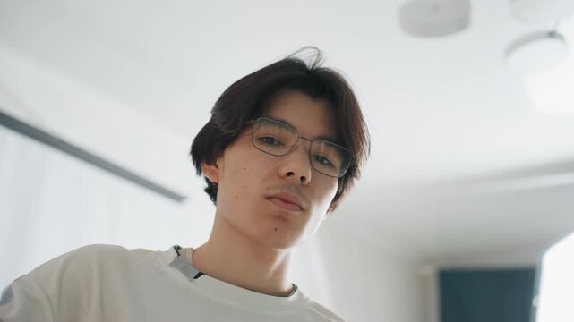 Male Asian person in living space assessing ceiling light with relaxed demeanor. Young Asian male examines ceiling illumination calmly within minimalist apartment setting prior to cleaning