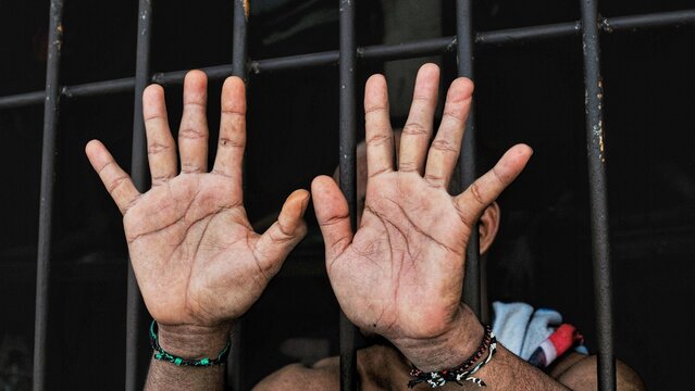 Close up of open palms of an inmate against iron bars of a prison cell, representing surrender, innocence, human rights, and plea for freedom
