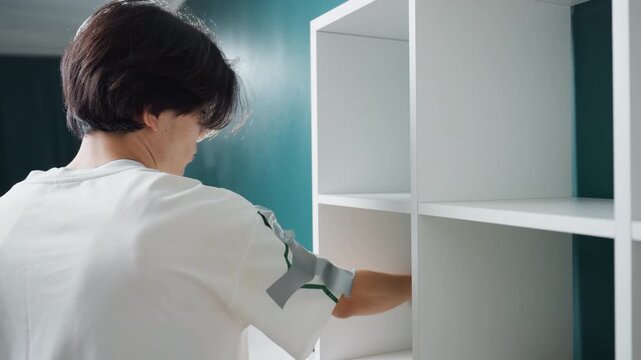 Youthful person cleaning and arranging simple shelves with care. An energetic young person diligently cleans and arranges neatly white shelves using spray and cloth in minimalist setting