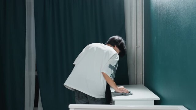 Methodical cleaning of household cabinet using spray and cloth. Diligent young person performs detailed disinfecting routine on home cabinet surface with spray and microfiber cloth