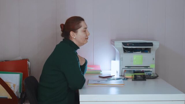 Woman holding throat while dialing from home desk, seeking telehealth consultation for shortness of breath, printer and notes visible, anxious expression, calling for medical advice