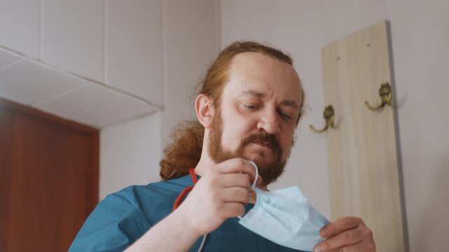 Paramedic donning mask in house hallway, bearded medic in scrubs adjusts mask and stethoscope, calm focused expression, warm interior light, coat hooks on wall, clinical readiness for home emergency
