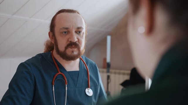 White paramedic home consultation calm attic setting with stethoscope, male clinician in blue scrubs speaks to patient, focused eye contact, note taking, supportive posture, residential lighting